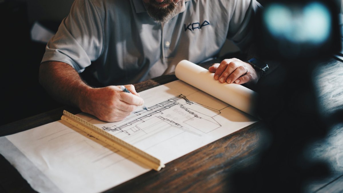Hands annotating a rolled architectural drawing with a pencil and a folding rule on a dark wooden desk.