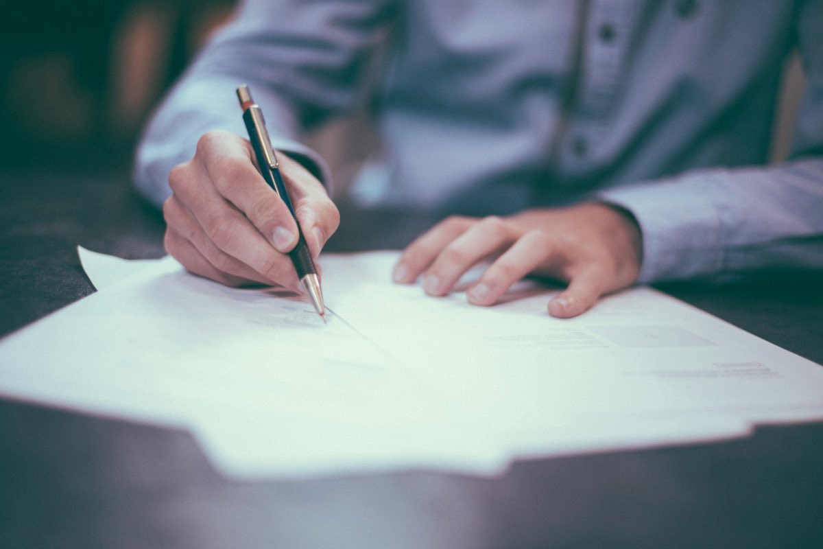 A pair of hands signing a printed document with a pen on a dark desk, lit from one side.