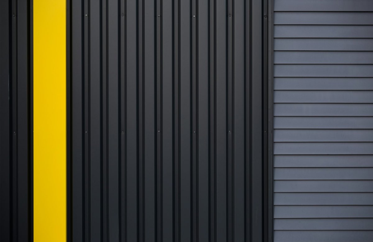 Dark ribbed metal cladding running vertically, interrupted by one bright painted band, beside a panel of grey horizontal siding.